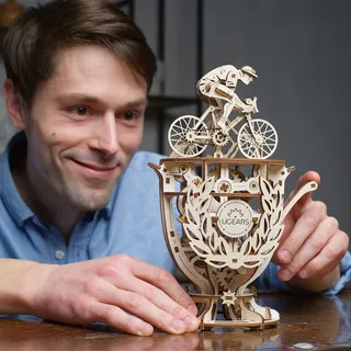 Un homme souriant, accoudé à une table, regarde le cycliste en bois sur sa coupe