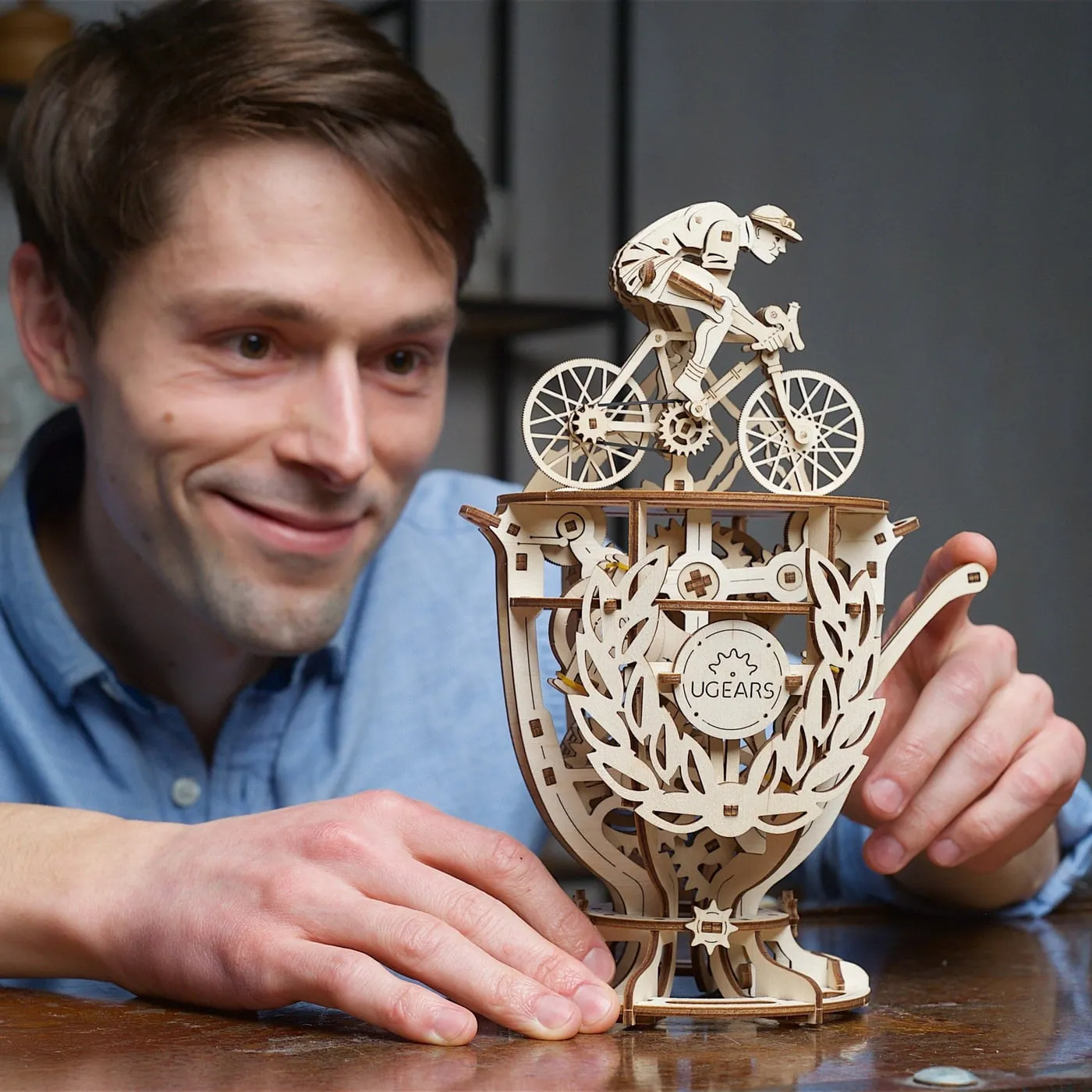 Un homme souriant, accoudé à une table, regarde le cycliste en bois sur sa coupe