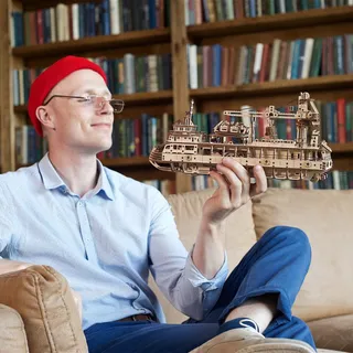 Un homme au bonnet rouge, assis dans un fauteuil, admire le bateau en bois qu’il tient à bout de bras