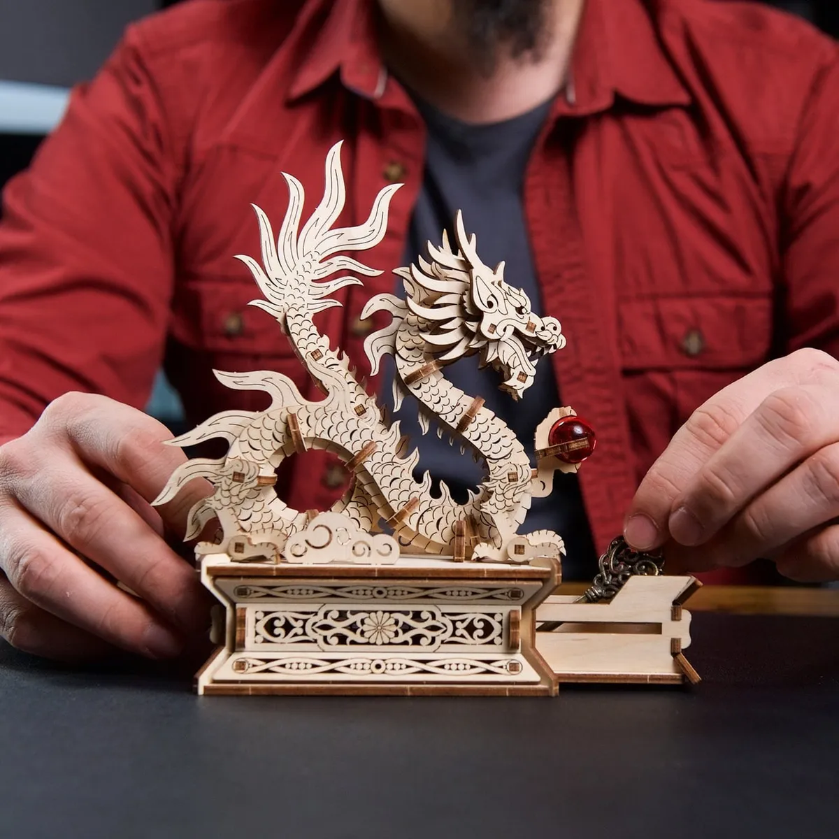 Un homme en chemise rouge ouvre le tiroir secret du socle, le dragon de bois dressé au-dessus