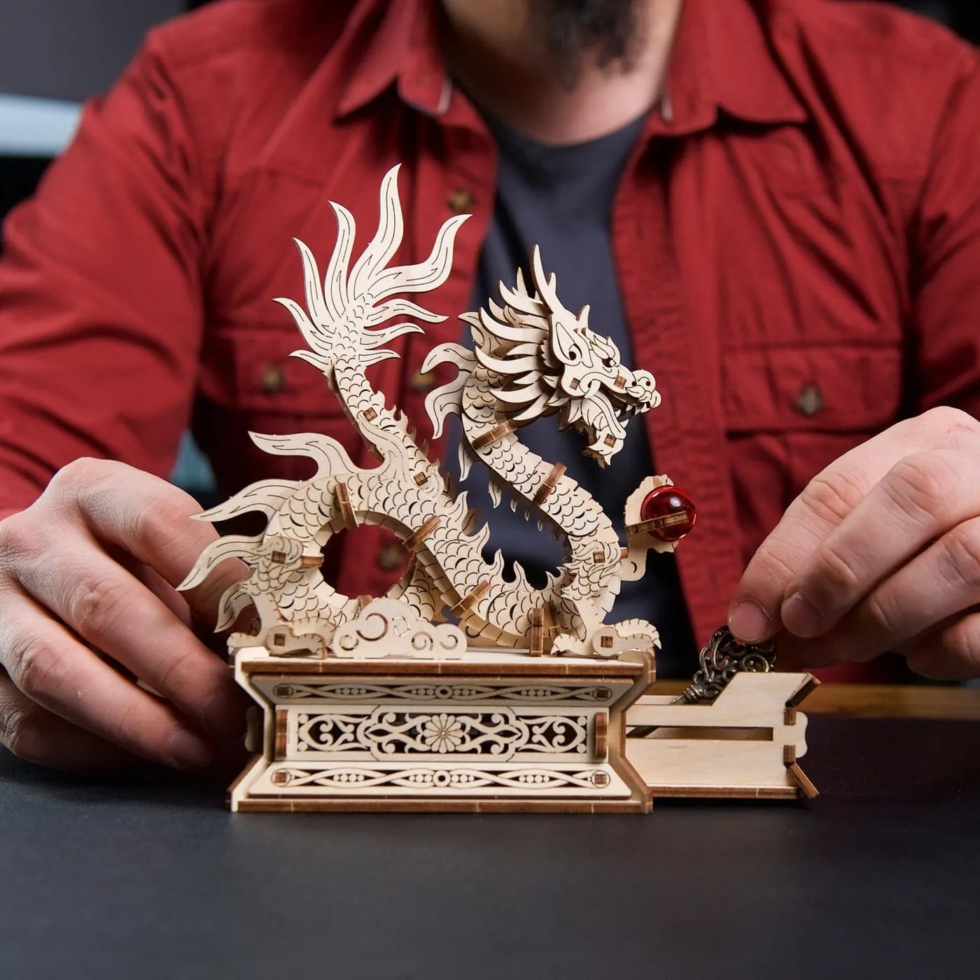 Un homme en chemise rouge ouvre le tiroir secret du socle, le dragon de bois dressé au-dessus