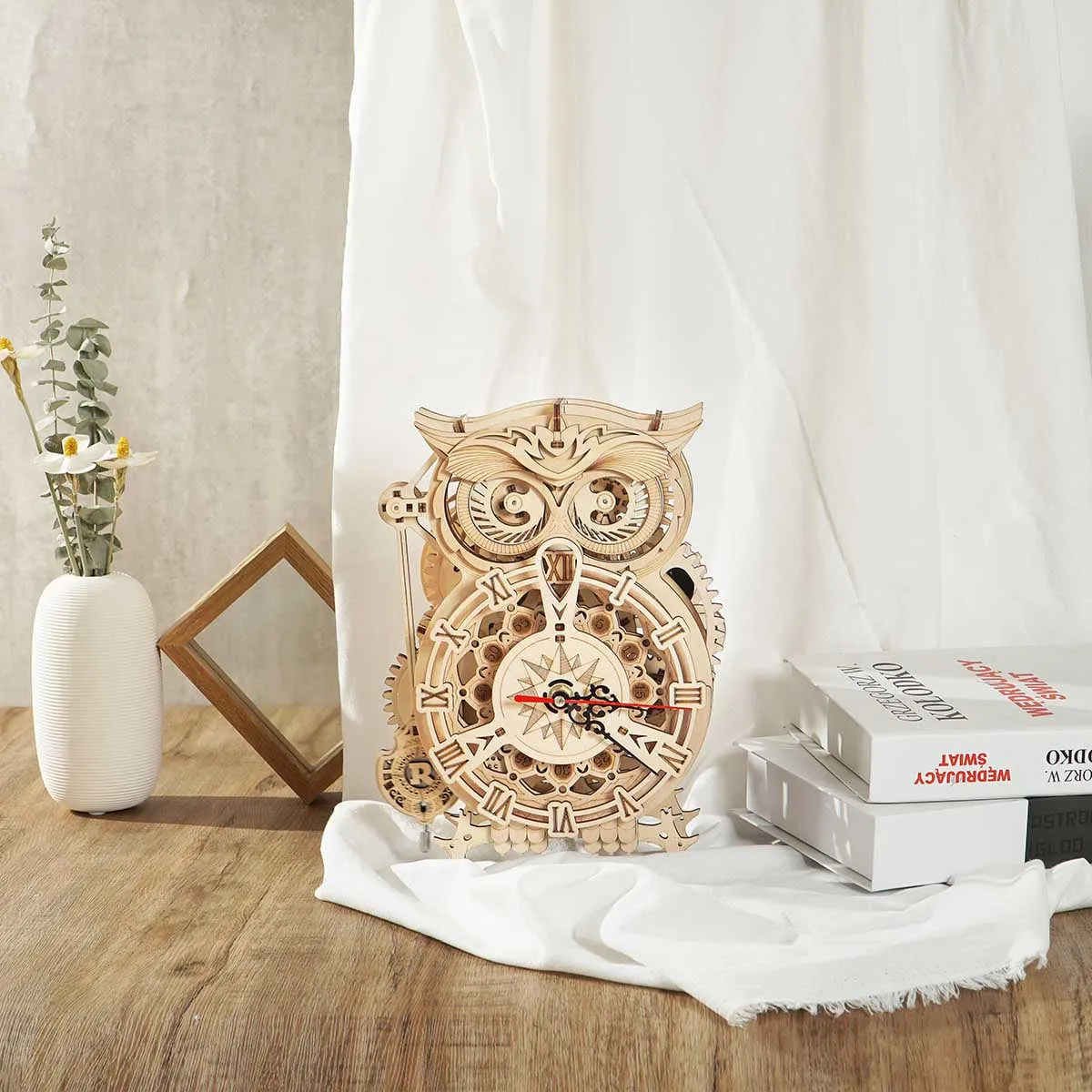 Horloge hibou sur une table en bois entre un vase de fleurs séchées, un cadre et des livres