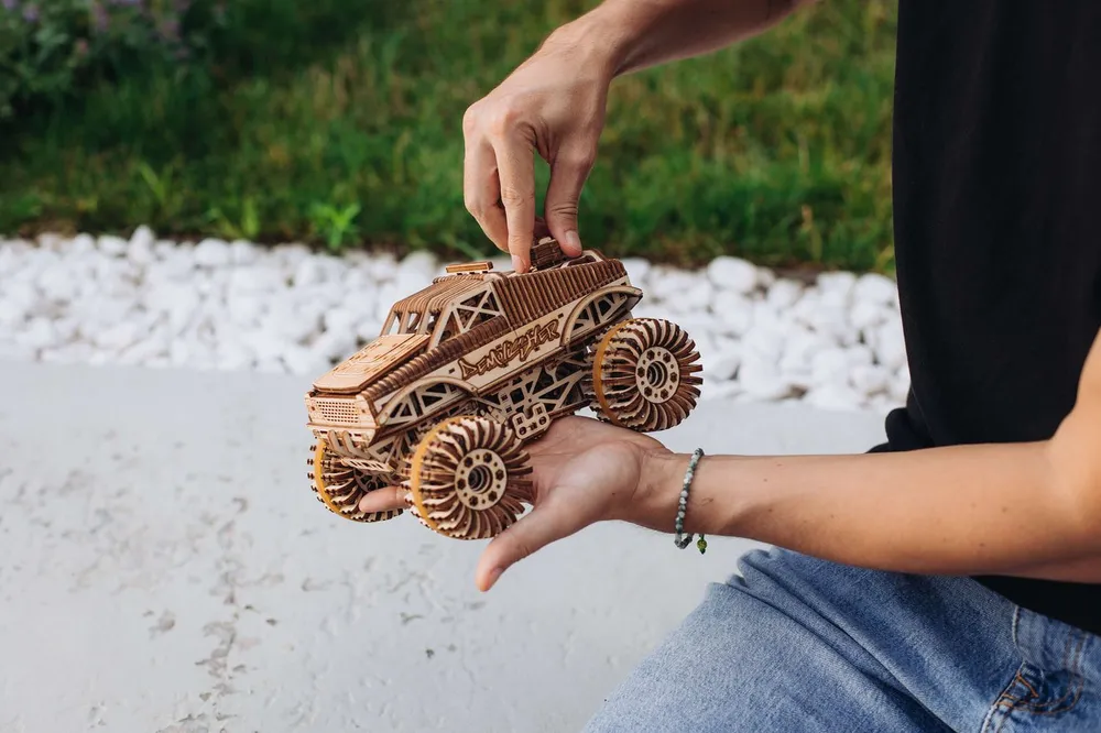 Une main pose le monster truck en bois sur un muret, devant des galets blancs