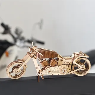 La moto en bois sur une table sombre, béquille centrale dépliée, devant une vraie moto floue à l’arrière-plan