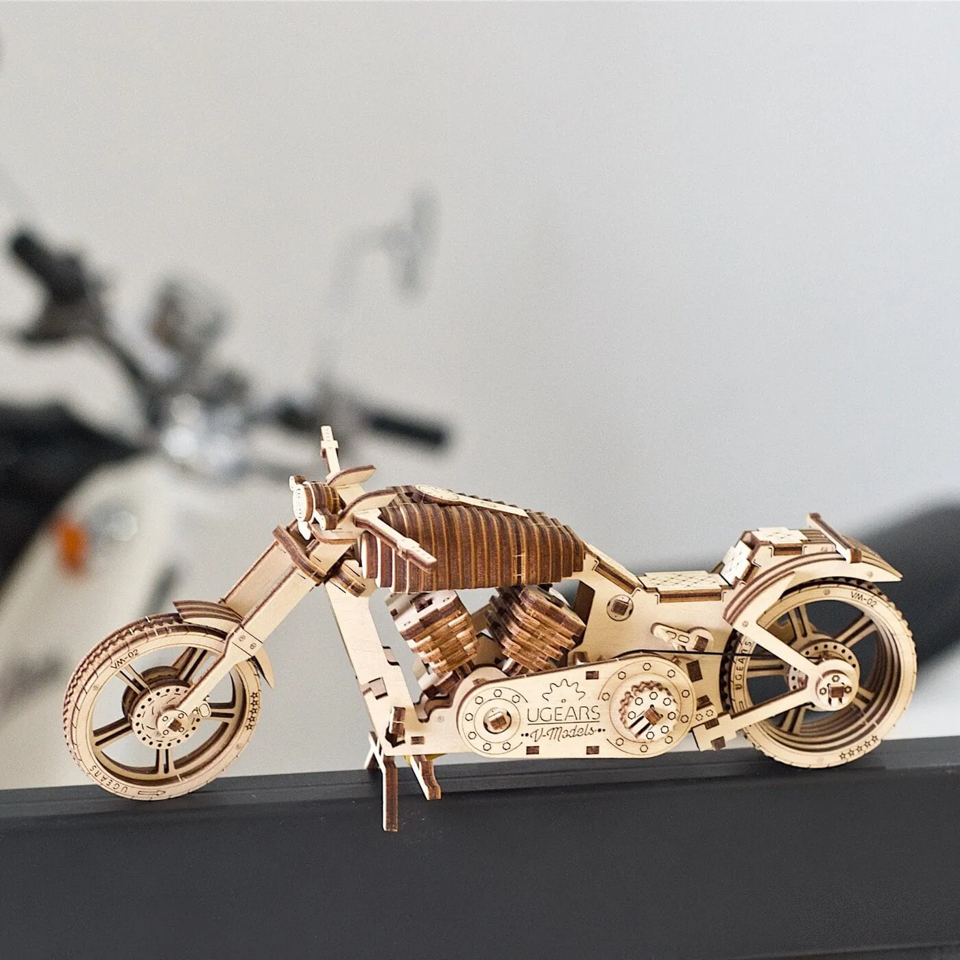 La moto en bois sur une table sombre, béquille centrale dépliée, devant une vraie moto floue à l’arrière-plan