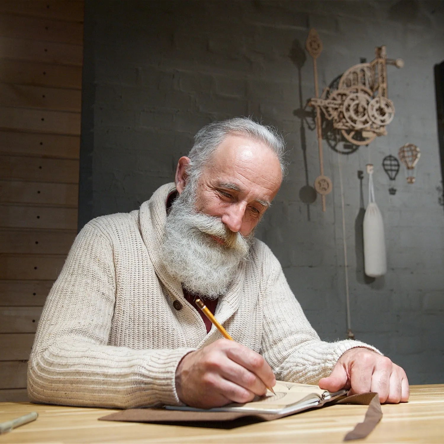 Un artisan barbu dessine à son établi, l'horloge en bois accrochée au mur derrière lui
